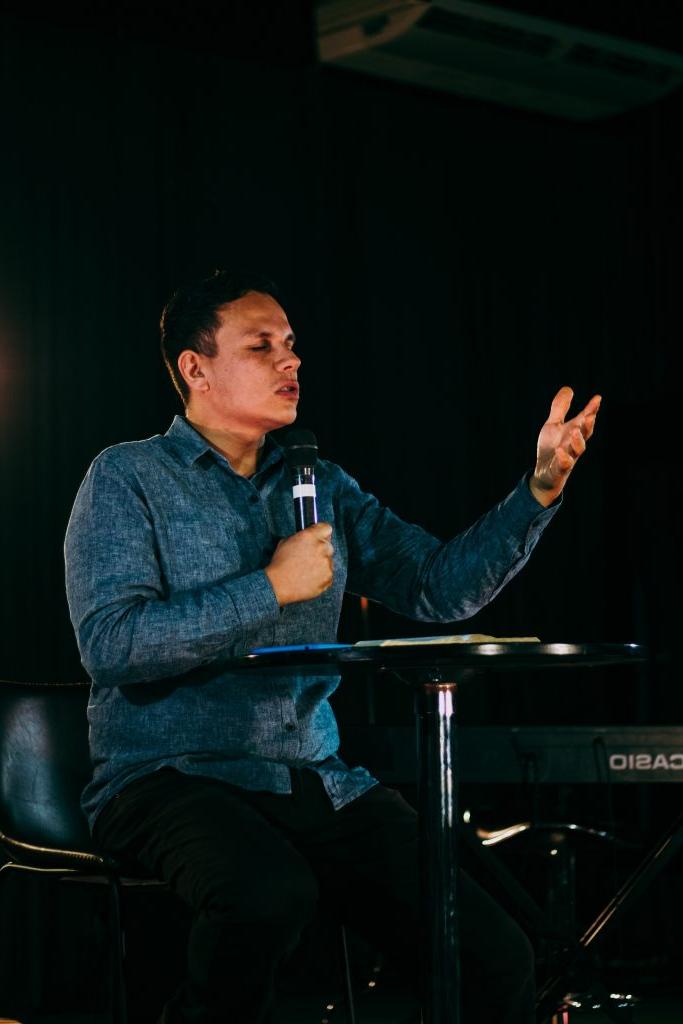 Young man sitting at small round table holding a microphone and praying.